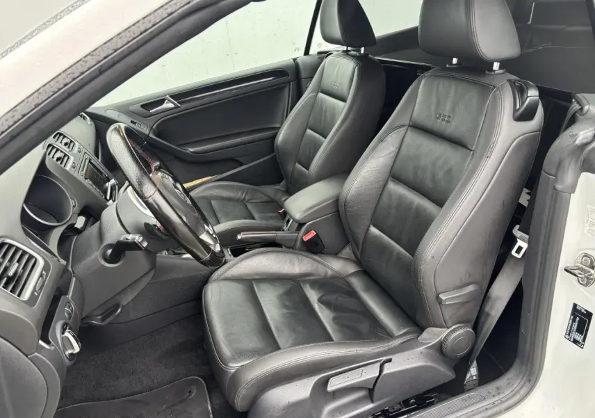 Vue intérieure côté conducteur de la Volkswagen Golf VI Cabriolet blanche, sièges sport en cuir noir avec logo GTI brodé.