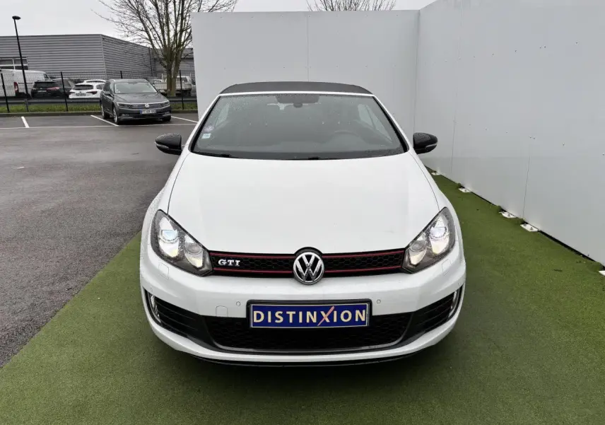 Vue avant d'une Volkswagen Golf VI Cabriolet GTI blanche avec calandre nid d'abeille et logo GTI rouge.