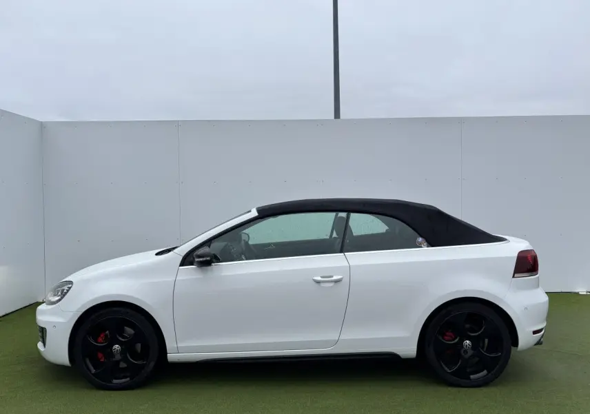 Profil côté gauche d'un Volkswagen Golf VI Cabriolet blanc avec toit souple noir et jantes alliage noires.