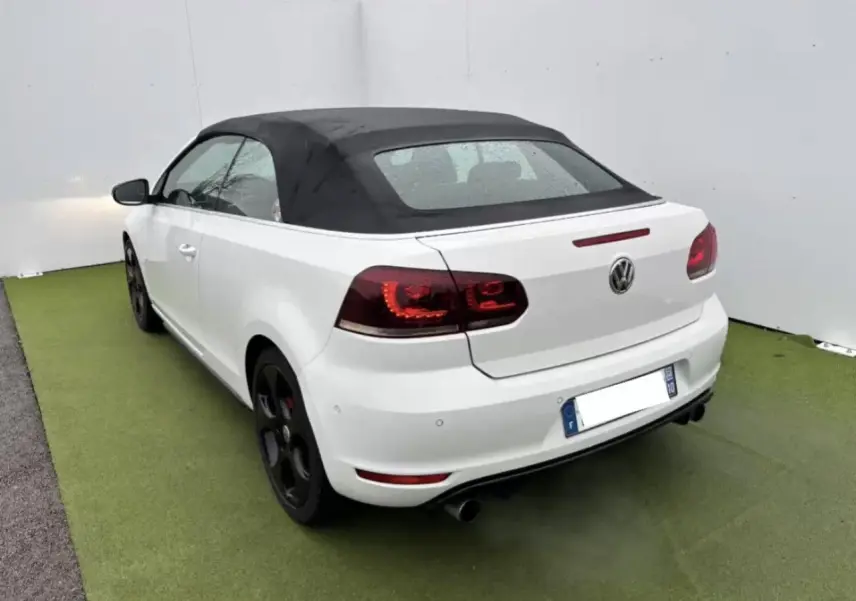 Vue 3/4 arrière droite d'une Volkswagen Golf VI Cabriolet blanche avec toit souple noir et jantes alliage foncé.
