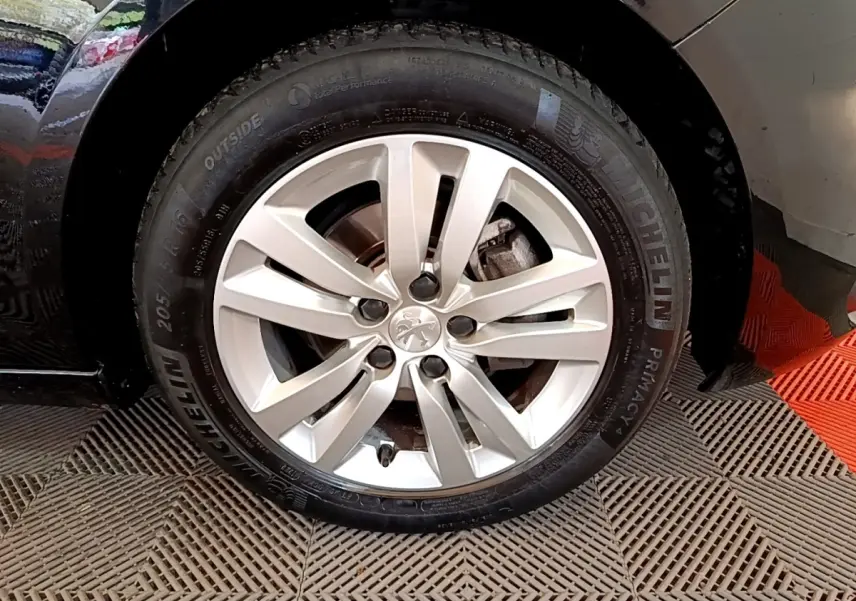 Gros plan sur la roue avant droite d’une Peugeot 308 noire, jante alliage argentée avec logo lion central.