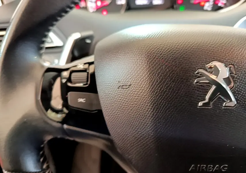 Gros plan sur le volant cuir noir de la Peugeot 308 avec logo lion et commandes intégrées, tableau de bord flou en arrière-plan.