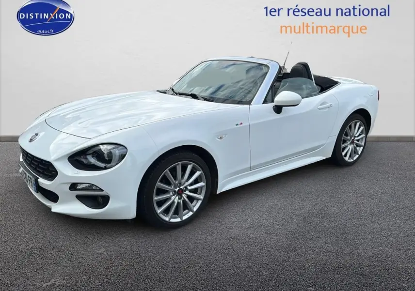 Fiat 124 Spider blanc vue 3/4 avant droit, capote ouverte, jantes alliage 17 pouces et phares antibrouillard visibles.