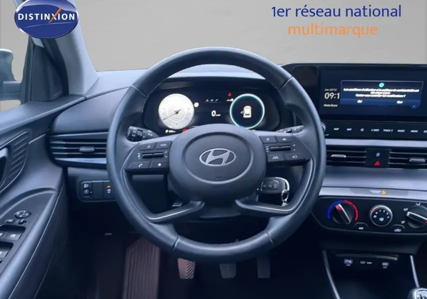 Vue intérieure centrée sur le volant cuir et tableau de bord numérique de la Hyundai i20 blanche, avec commandes au volant visibles.