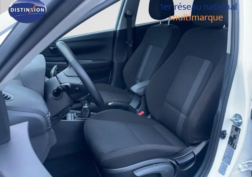 Vue intérieure côté conducteur d'une Hyundai i20 blanche, montrant les sièges noirs en tissu et la boîte manuelle.