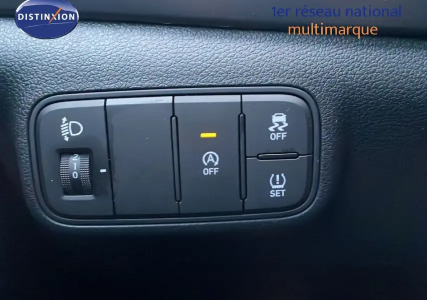 Gros plan sur les boutons de contrôle noirs du tableau de bord d’une Hyundai i20 blanche, avec réglage de phares et aides à la conduite.