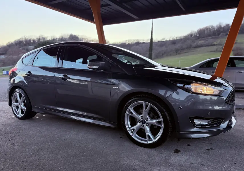 Ford Focus 1.5 TDCI ST Line grise vue de profil côté gauche sous abri, avec jantes alliage 18 pouces et phares allumés.