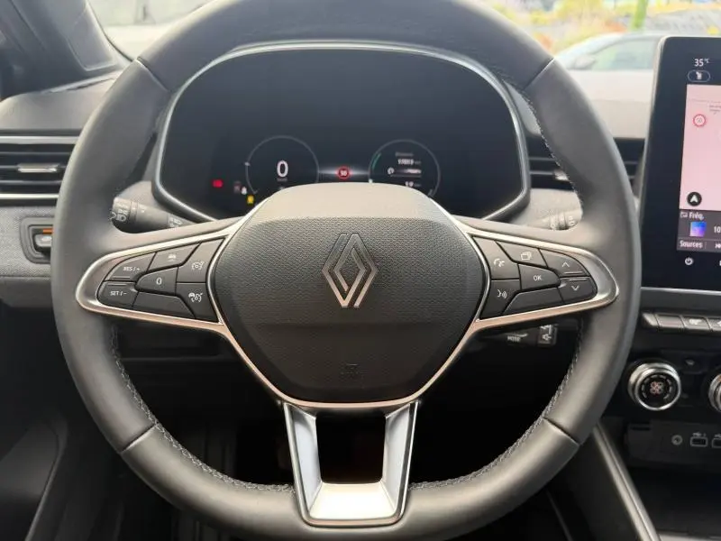 Vue rapprochée du volant multifonction noir avec logo Renault au centre, tableau de bord numérique en arrière-plan.