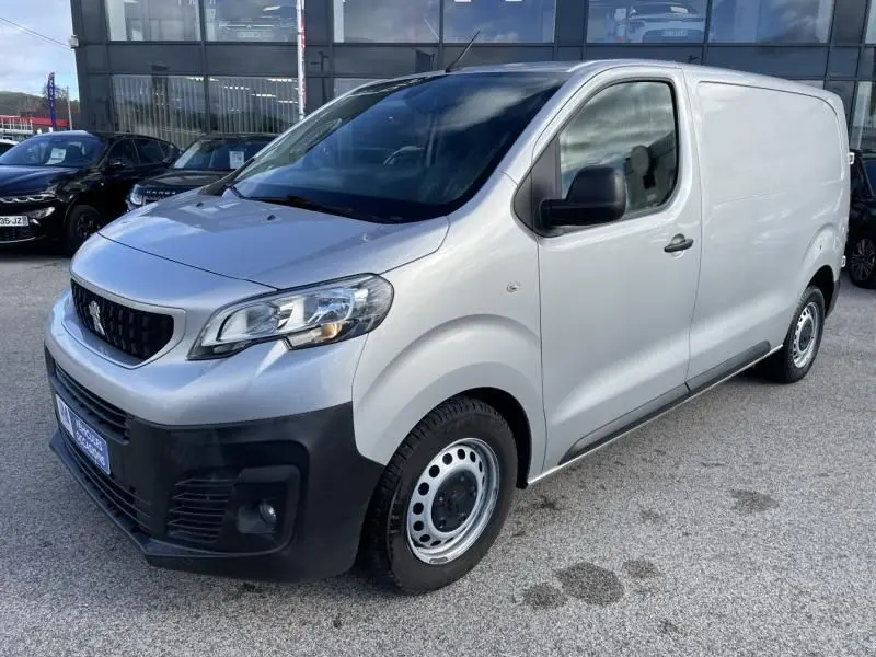 Vue 3/4 avant droit d’un Peugeot Expert Fourgon gris métal avec pare-chocs noir et jantes acier, stationné devant un bâtiment vitré.