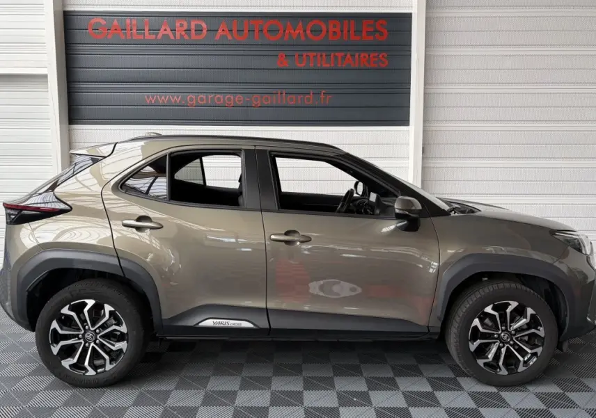 Profil droit d'une Toyota Yaris Cross 2024 gris clair avec jantes alliage noires et logo Yaris Cross sur la porte.