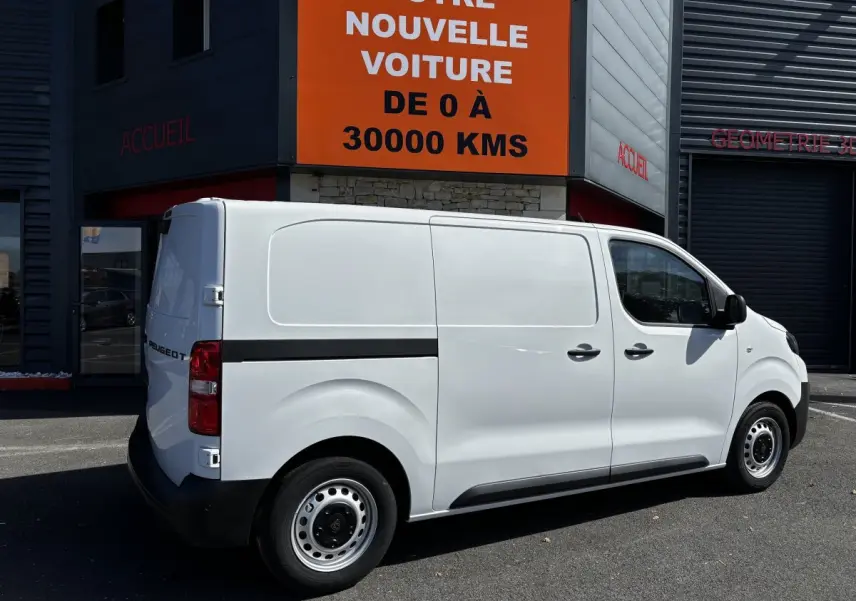 Peugeot Expert Fourgon blanc vu de profil côté droit, stationné devant un bâtiment commercial avec panneau orange.