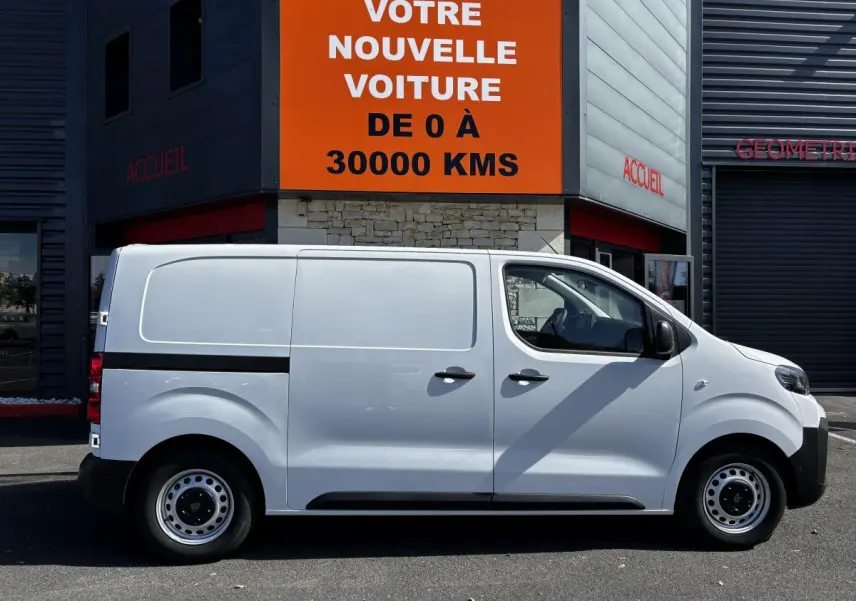 Profil côté droit du Peugeot Expert Fourgon blanc 2025 garé devant un bâtiment avec panneau orange.