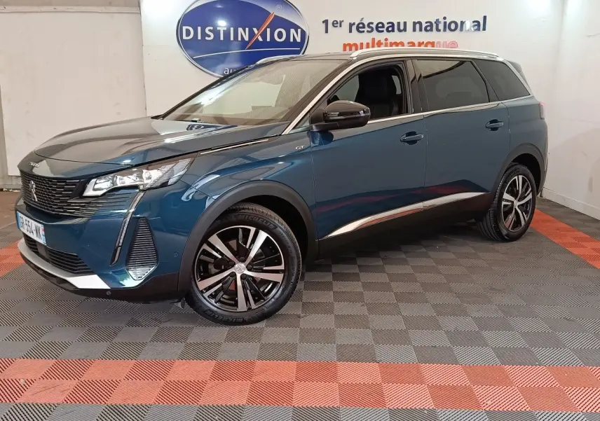 Peugeot 5008 bleu en 3/4 avant droit, avec jantes noires et détails chromés, sur sol damier intérieur showroom.