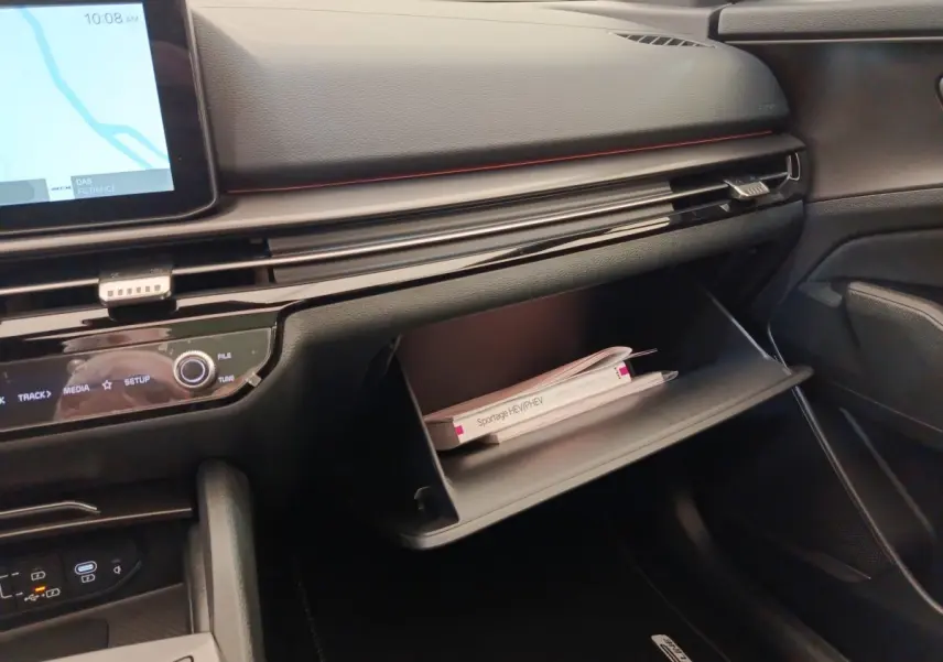 Intérieur KIA Sportage 2026, vue côté passager sur tableau de bord noir avec boîte à gants ouverte et manuel visible.