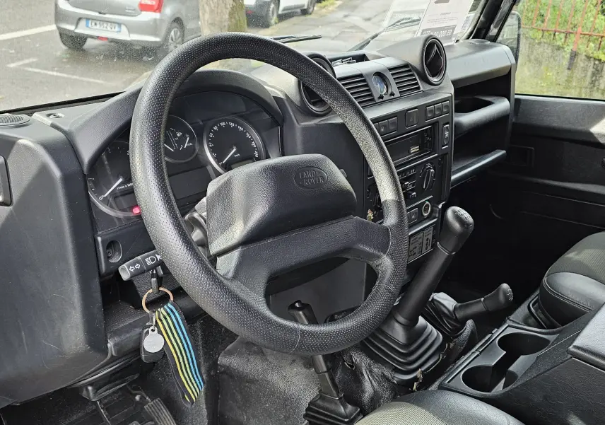 Intérieur noir du Land Rover Defender 90 2013, vue côté conducteur sur le volant, levier de vitesses manuel et tableau de bord.