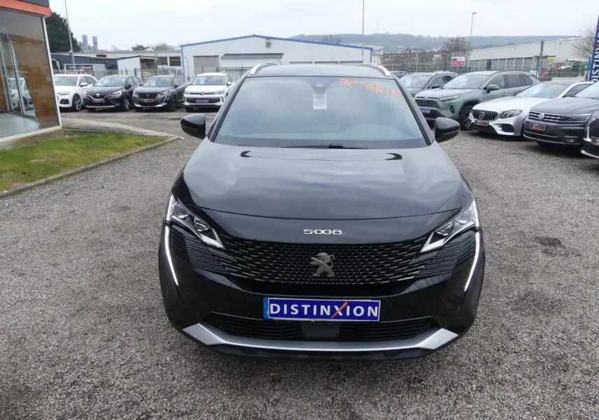 Vue frontale d'un Peugeot 5008 noir métallisé avec calandre GT et feux LED verticaux allumés sur un parking.