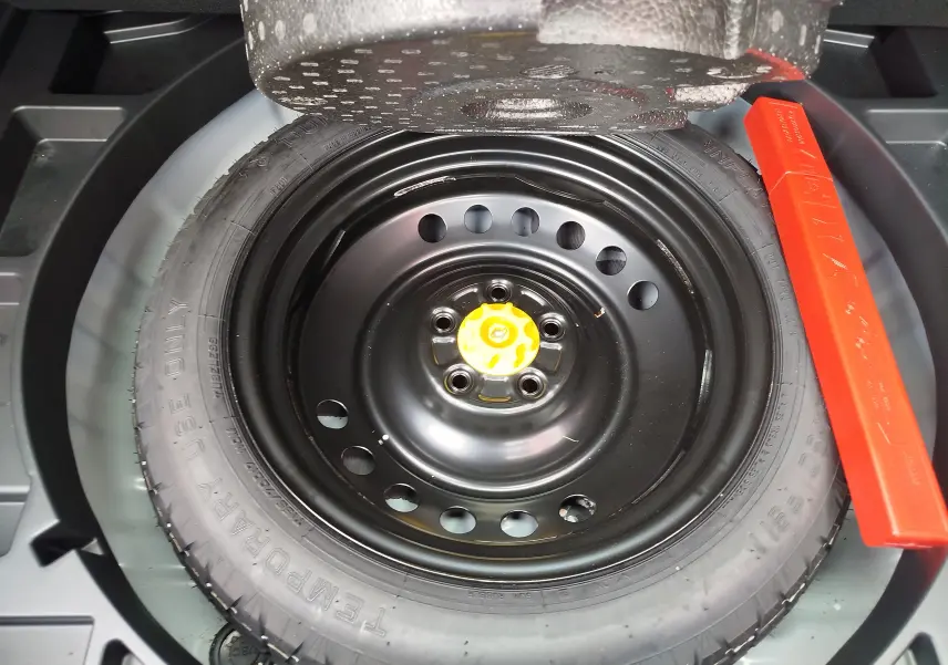 Vue rapprochée de la roue de secours noire avec écrou jaune et triangle de signalisation rouge dans le coffre d'une Toyota Corolla Cross.