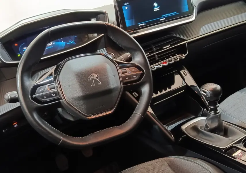 Vue intérieure centrée sur le volant cuir et la console centrale avec levier de vitesse manuelle de la Peugeot 208 blanche.