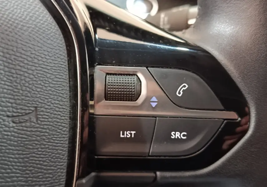 Gros plan sur les commandes noires du volant côté gauche d'une Peugeot 208 blanche, version PureTech 100 S&S Allure Business.