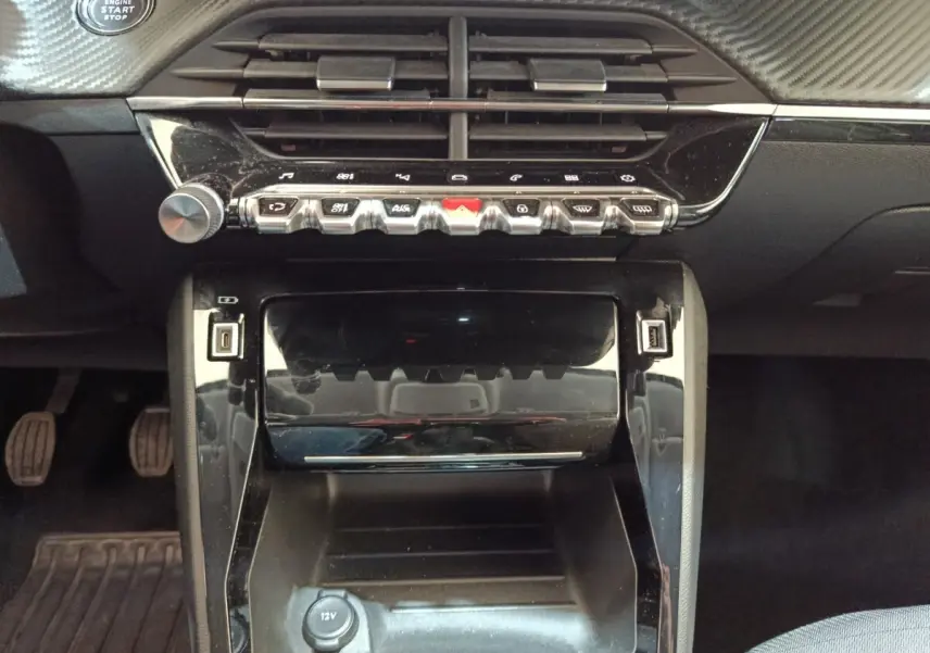 Vue rapprochée du tableau de bord central noir brillant de la Peugeot 208 PureTech 100, avec commandes et prises USB visibles.