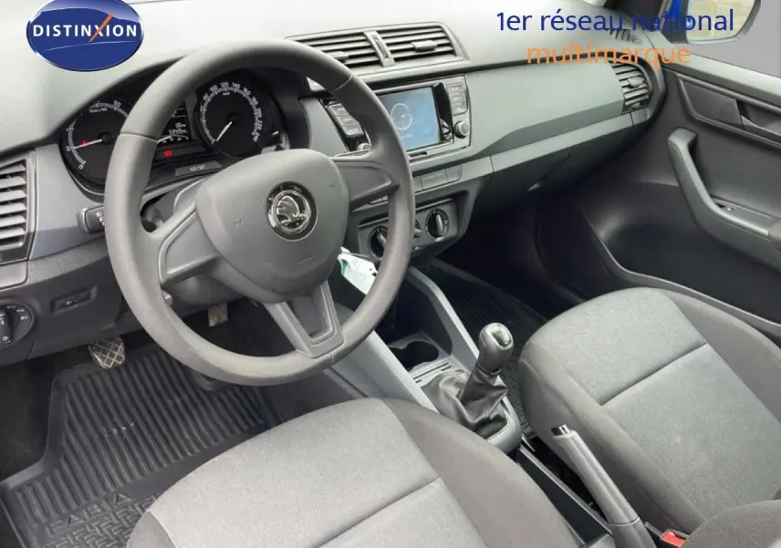 Vue intérieure côté conducteur de la Skoda Fabia 2019, tableau de bord noir et sièges tissu gris, volant et levier de vitesse visibles.