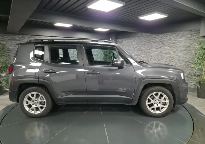 Profil côté gauche d'un Jeep Renegade gris métallisé avec toit noir et jantes alliage 17 pouces en intérieur showroom.