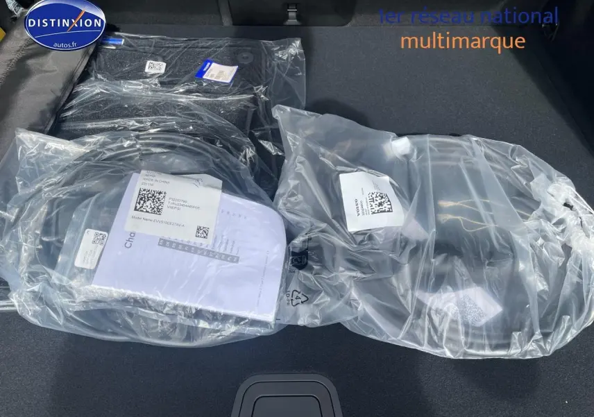 Coffre ouvert d'un Volvo XC60 gris 2024, montrant tapis et accessoires neufs emballés en plastique transparent.