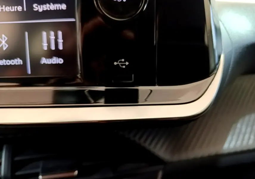 Gros plan sur la console centrale noire brillante de la Peugeot 208 blanche, avec port USB et écran tactile partiel visible.