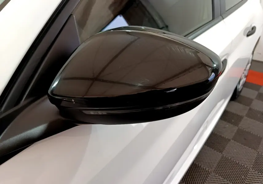 Gros plan sur le rétroviseur noir côté gauche d'une Peugeot 208 blanche vue de profil, stationnée en intérieur.