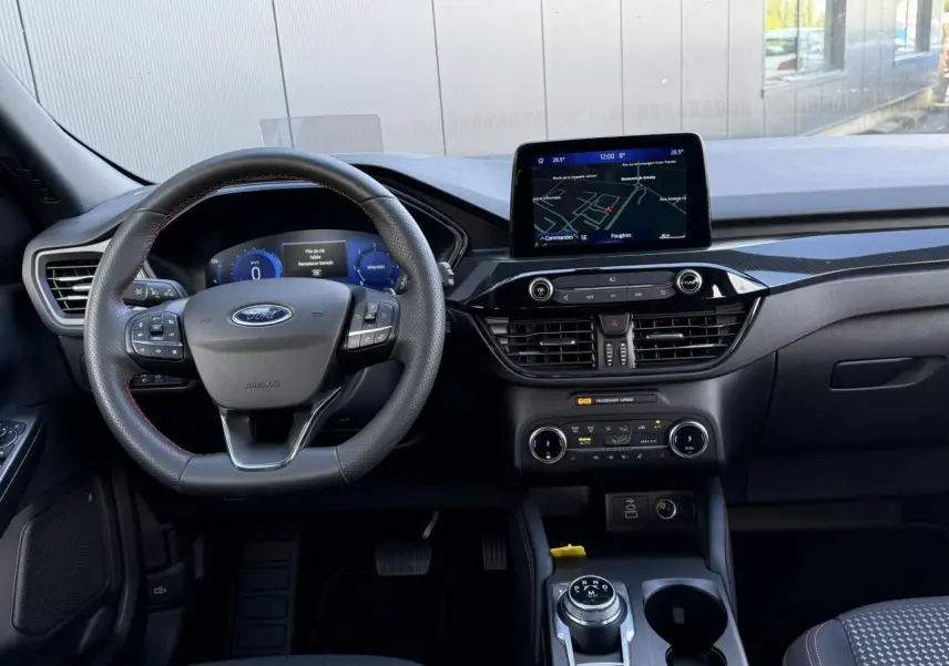 Vue intérieure avant du Ford Kuga 2024 ST-Line, tableau de bord noir avec écran tactile et volant à méplat avec surpiqûres rouges.