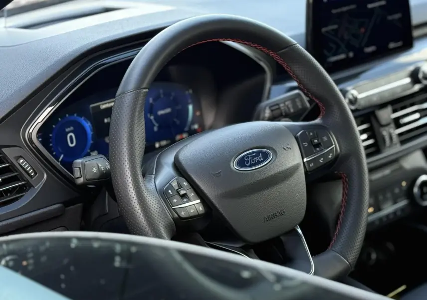 Vue rapprochée du volant noir avec surpiqûres rouges du Ford Kuga 2024 ST-Line, tableau de bord numérique en arrière-plan.