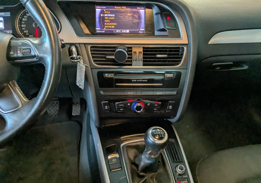 Vue intérieure centrée sur la console centrale et levier de vitesse manuelle d'une Audi A4 gris 2013 avec écran multimédia allumé.