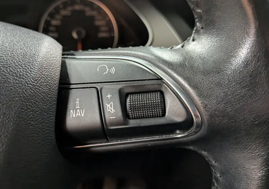 Gros plan sur les commandes de navigation et volume sur le volant en cuir noir d'une Audi A4 gris diesel de 2013.