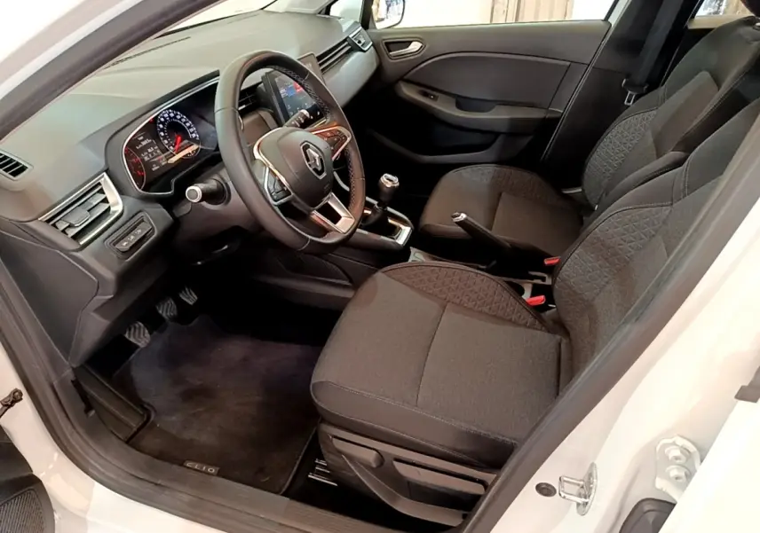 Vue intérieure côté conducteur d'une Renault Clio Business blanche, montrant sièges noirs et tableau de bord avec volant multifonctions.
