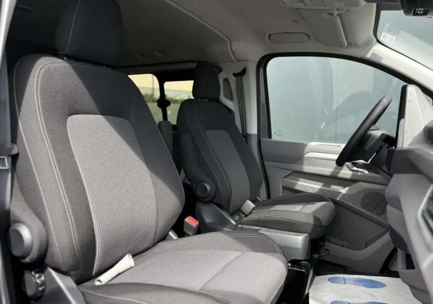 Vue intérieure côté conducteur du Volkswagen Transporter T7 ProCab 2026, sièges tissu gris à rayures et volant multifonction.
