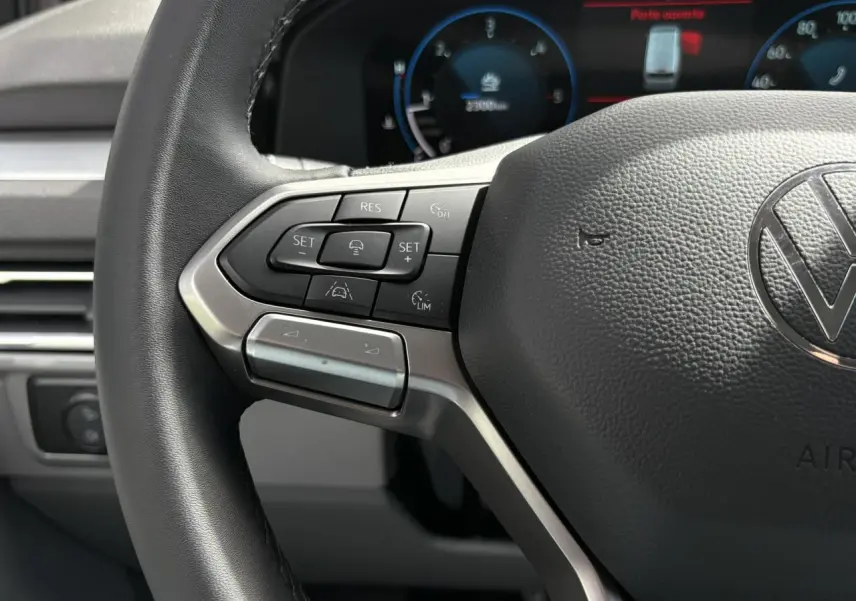 Gros plan sur le volant multifonction noir du Volkswagen Transporter T7 avec commandes de régulateur de vitesse et tableau de bord numérique en arrière-plan.