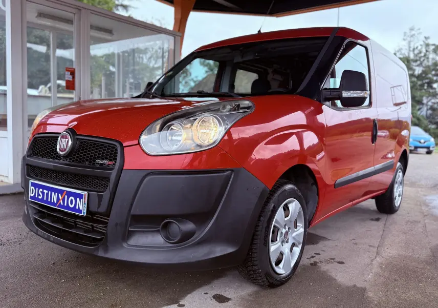 Vue 3/4 avant droit d’un FIAT Doblo Cargo rouge avec pare-chocs noirs et jantes argentées sous abri.