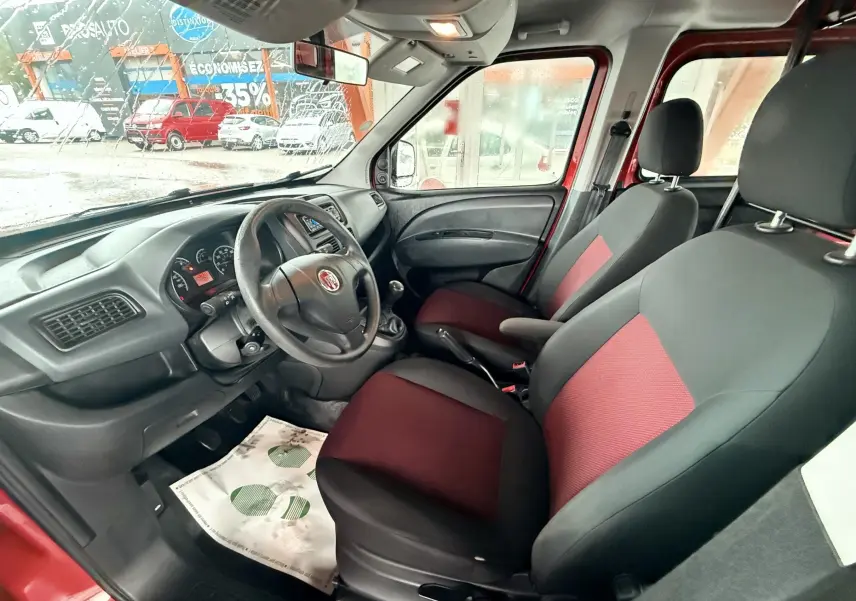 Intérieur du Fiat Doblo Cargo 2011 vu côté conducteur, sièges tissu rouge et noir, volant cuir et tableau de bord noir.