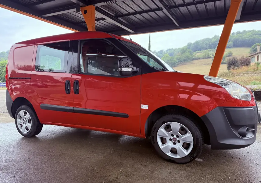 Fiat Doblo Cargo rouge vu de profil côté gauche, sous un abri avec arrière-plan de collines verdoyantes.