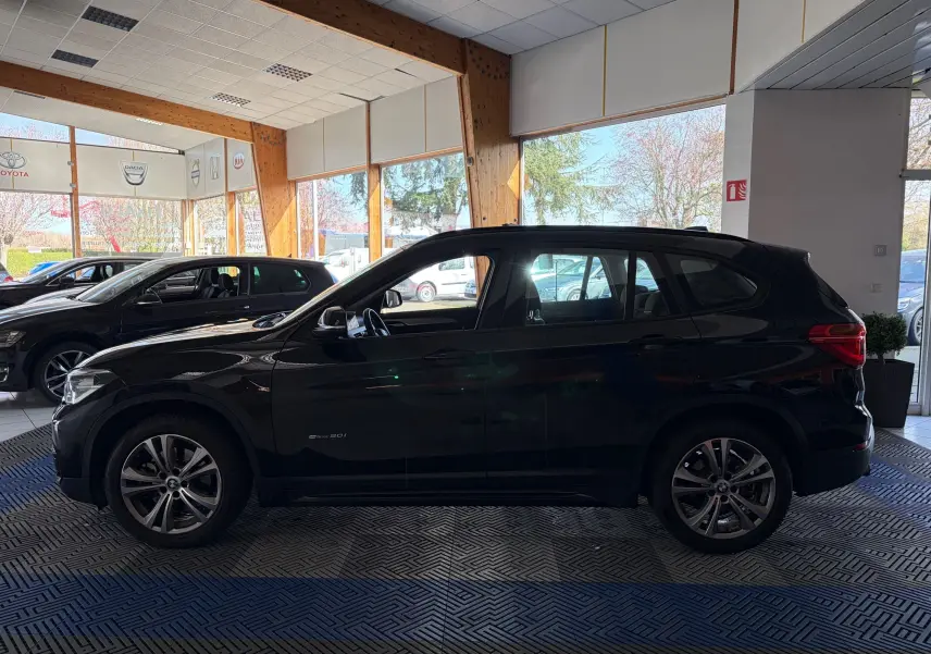 BMW X1 20i S-Drive Sport Line noir vue de profil côté gauche en intérieur de showroom lumineux.
