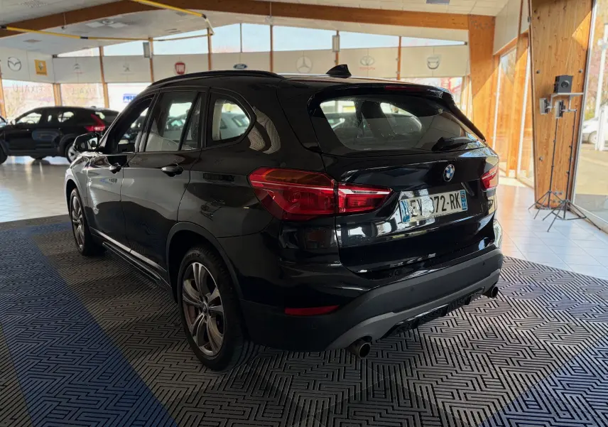 BMW X1 20i S-DRIVE noir vue 3/4 arrière droit en intérieur, avec feux arrière rouges et jantes alliage.