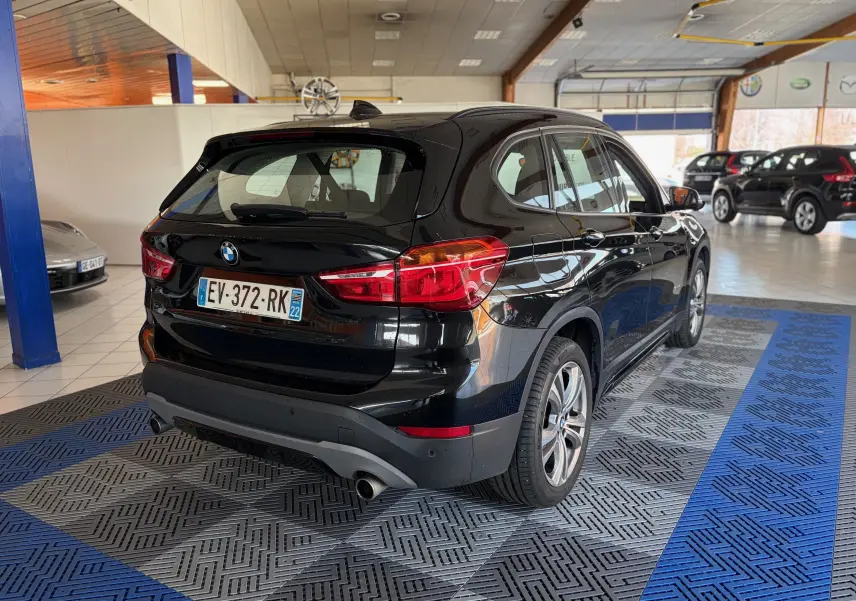 BMW X1 20i S-Drive Sport Line noir vue 3/4 arrière droit en intérieur avec feux arrière allumés et jantes alliage.
