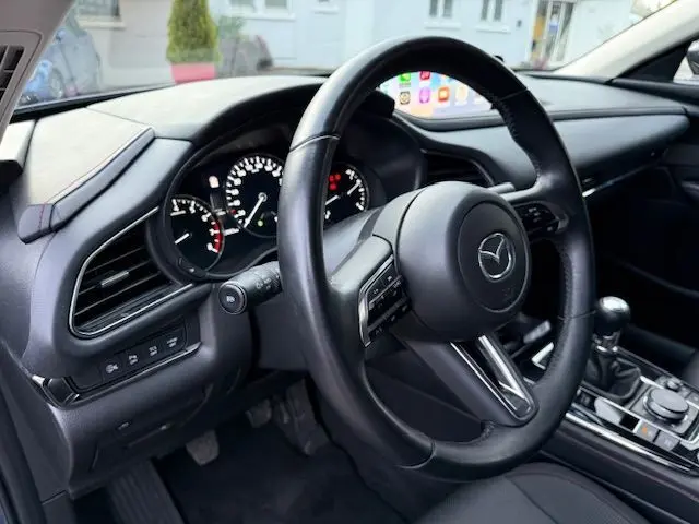 Vue intérieure côté conducteur du Mazda CX-30 gris, volant cuir avec commandes intégrées et levier de vitesse manuel visible.