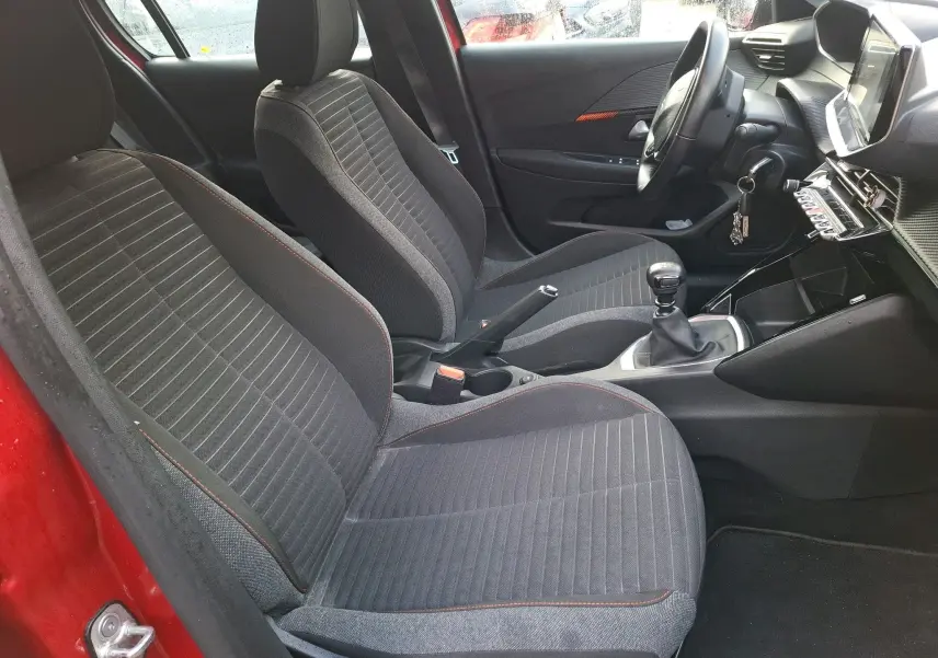 Vue intérieure côté gauche de la Peugeot 208 rouge Elixir, montrant les sièges avant gris avec surpiqûres orange et la boîte manuelle.