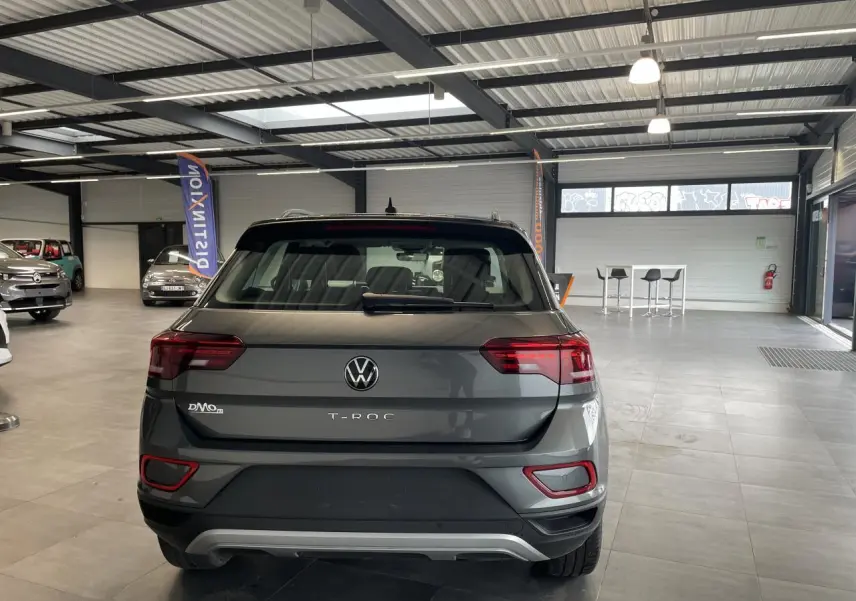 Vue arrière du Volkswagen T-Roc 2025 en gris Indium métallisé avec feux LED et diffuseur argenté dans un showroom.
