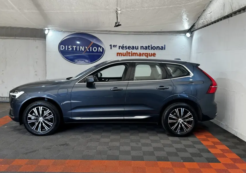 Profil droit d’un Volvo XC60 bleu hybride rechargeable avec jantes alliage et rails de toit chromés, en intérieur showroom.
