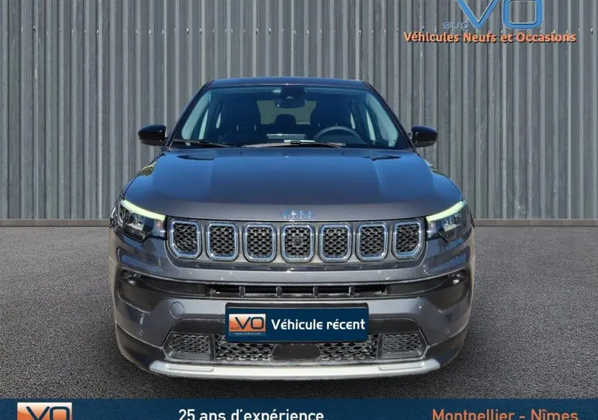 Vue frontale d'un Jeep Compass gris 2024 avec calandre distinctive et phares LED allumés sur fond industriel.
