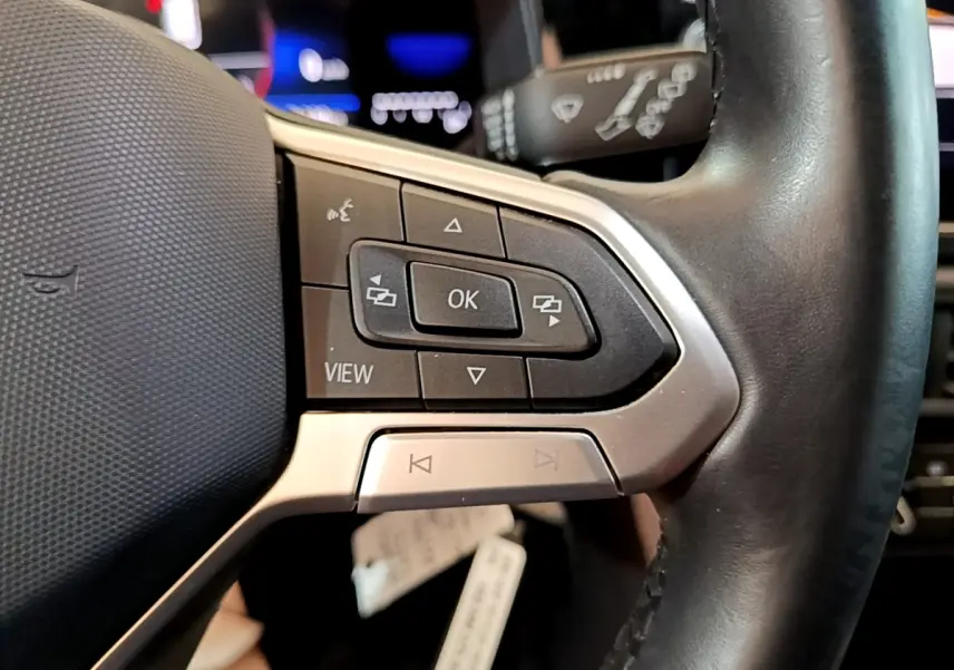 Gros plan sur les commandes gauche du volant cuir alu du Volkswagen Taigo noir, avec boutons multifonctions et affichage numérique flou.