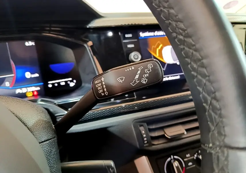 Gros plan sur la commande d’essuie-glace à gauche du volant cuir du Volkswagen Taigo noir, tableau de bord numérique en arrière-plan.