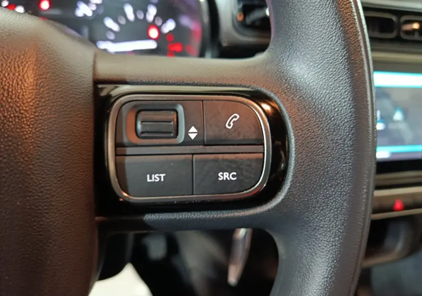 Gros plan sur les commandes gauche du volant noir d’une Citroën C3 2024, avec tableau de bord et écran tactile en arrière-plan.